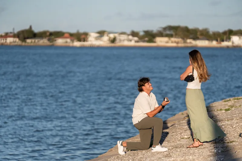 Michael kneels on the beach sand holding a ring box while Katie stands facing him with the ocean and coastal homes behind them.