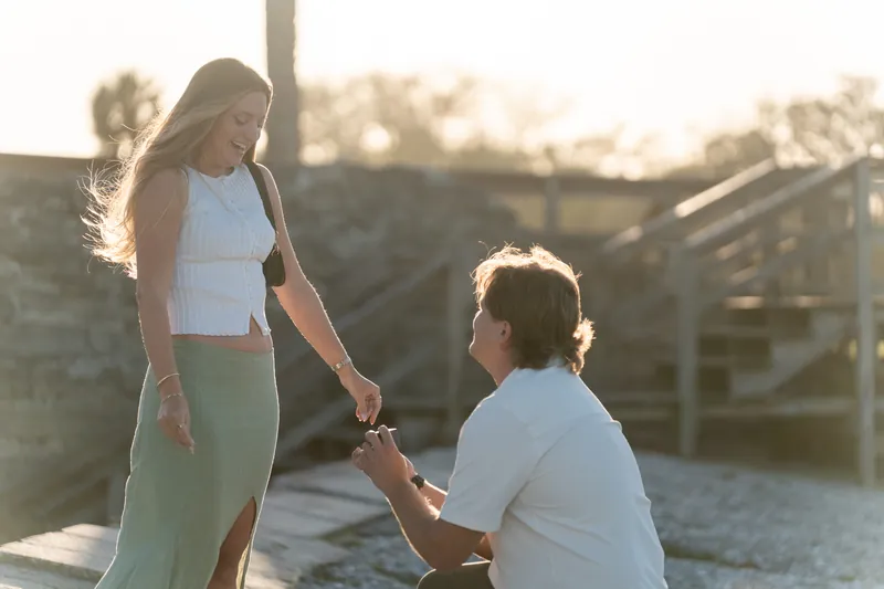Michael on one knee proposes to Katie on a wooden deck at sunset, holding her hand.