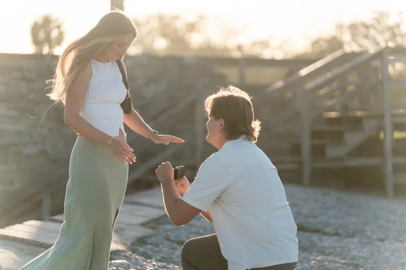 Katie looks down at Michael as he proposes on the deck with golden hour light surrounding them.