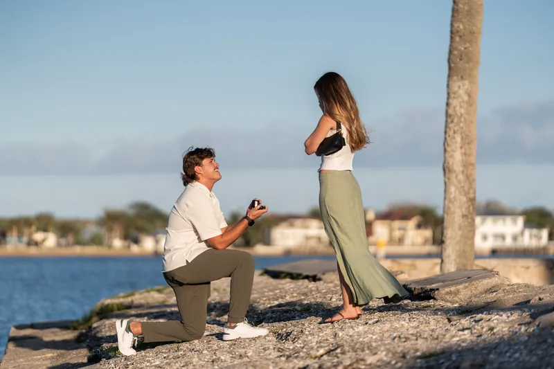 Michael holds Katie's hand while kneeling to propose on the beach near a palm tree.