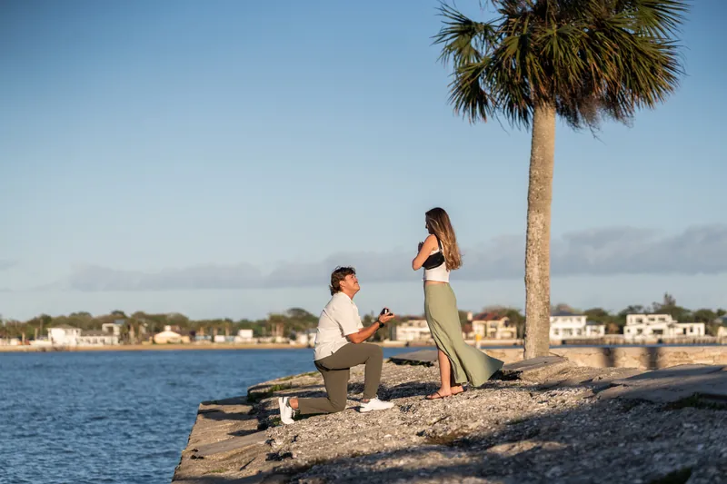 Michael kneels on a rocky beach pier holding a bouquet, proposing to Katie who stands before him with a palm tree and waterfront homes behind them.