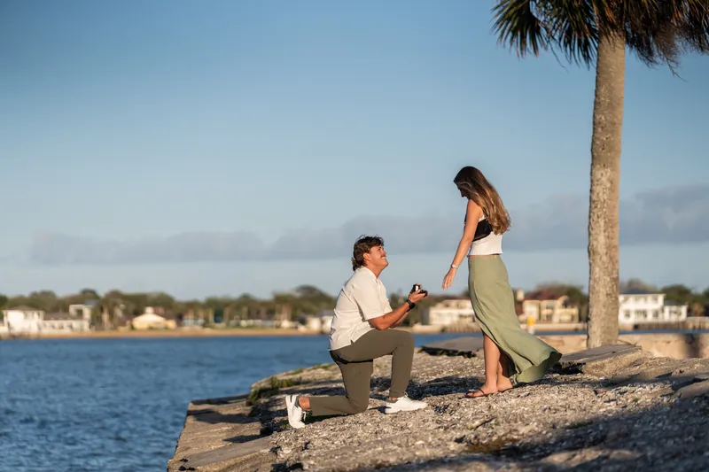 Michael presents an engagement ring to Katie as she reacts with surprise, standing on the sandy pier beneath a tall palm tree overlooking the bay.