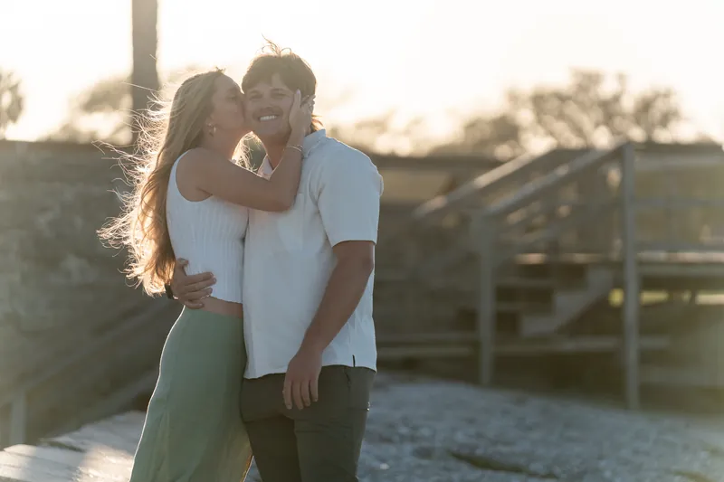 Katie hugs Michael from behind on a wooden dock at golden hour, both smiling as the sunset bathes them in warm light.