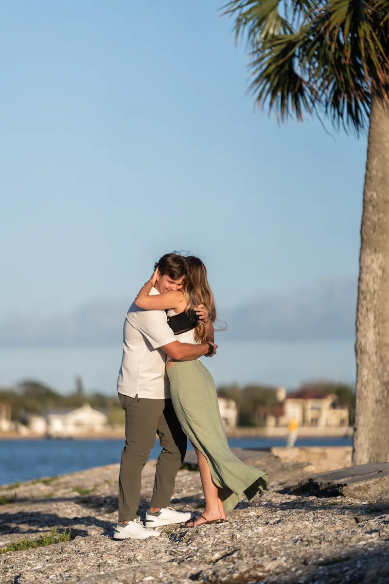 Michael and Katie embrace and kiss under a palm tree on the beach, the calm bay and coastal town visible across the water.