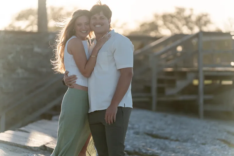 Katie rests her head on Michael's shoulder as they stand together on the dock, both wearing casual clothes with the waterfront and wooden railing behind them.