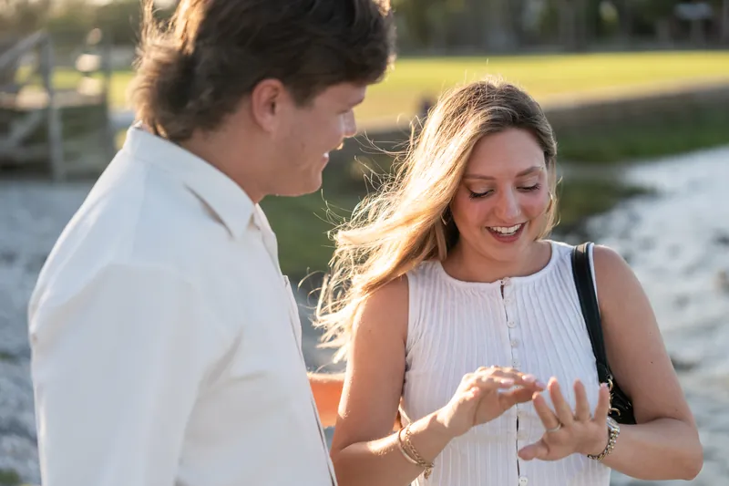 Michael slides an engagement ring onto Katie's finger as she smiles, with a waterfront and green grass behind them.