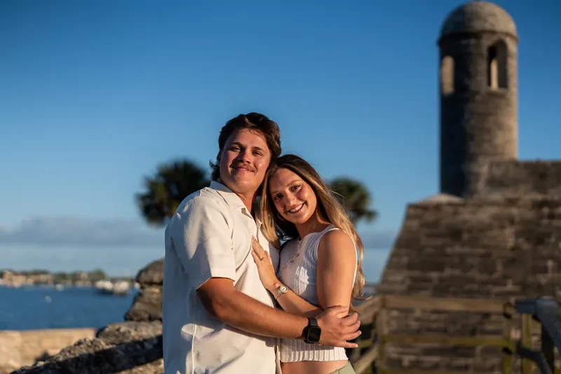 Michael and Katie embrace with a historic stone tower and blue water visible behind them under clear skies.