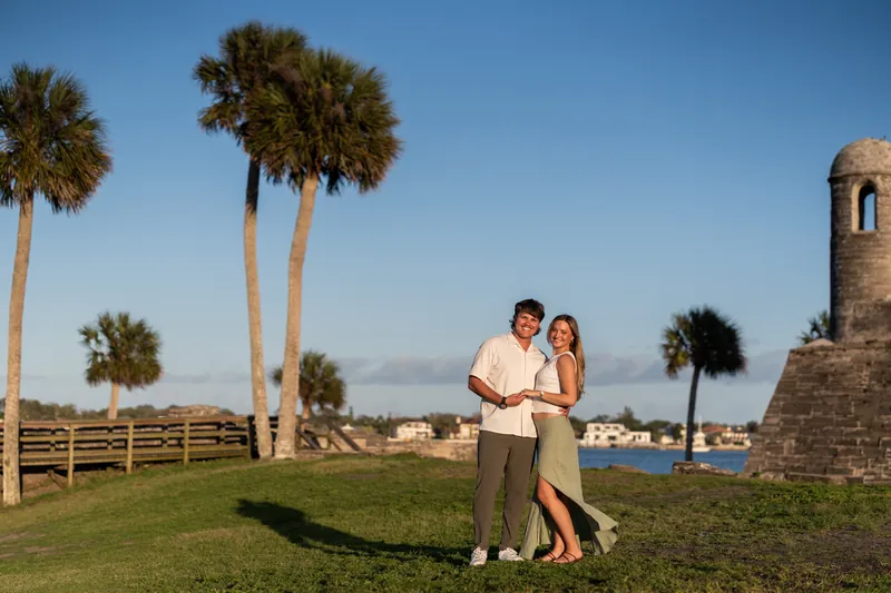 Michael and Katie stand in a grassy field lined with palm trees, a stone tower, and waterfront in the distance.