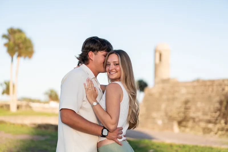 Michael kisses Katie's cheek as she smiles, wearing white and cream clothing with a stone tower and palm trees in soft focus behind them.