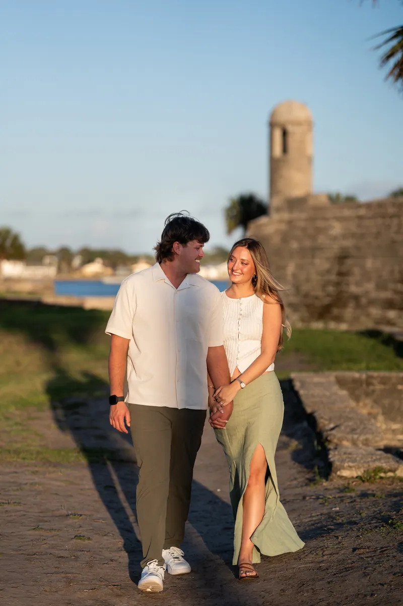 Michael and Katie walk hand-in-hand on a path with a historic stone tower, palm trees, and water visible in the background.