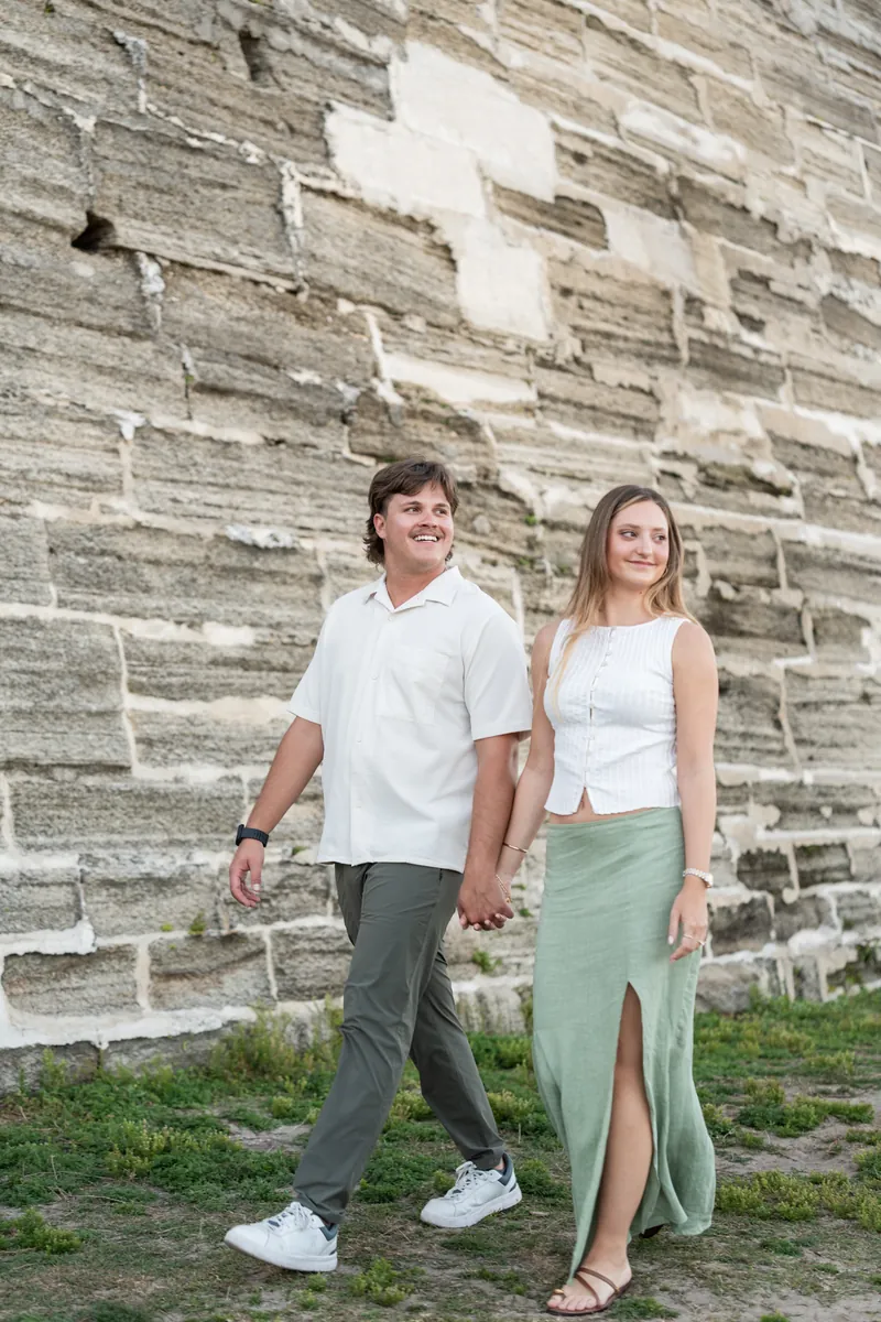 Michael and Katie hold hands while standing against a stone wall with a white cross, wearing casual white tops and neutral pants.