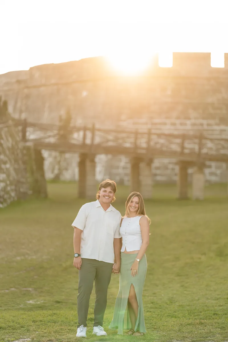 Michael and Katie stand together on a grassy field at sunset with a stone fortress and wooden bridge in the background.