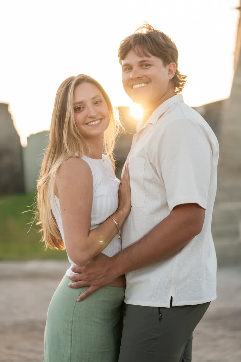 Michael and Katie embrace at golden hour, both wearing white sleeveless tops and green pants, with a blurred stone structure behind them.
