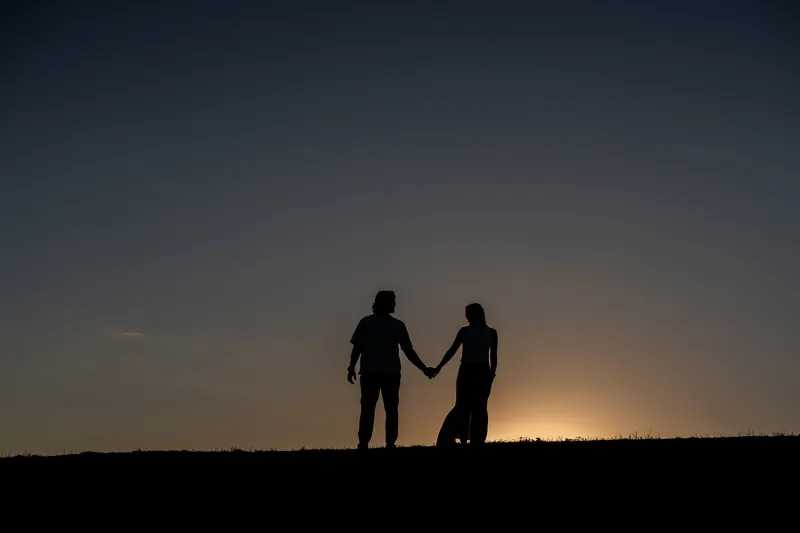 Silhouetted against a dramatic sunset, Michael and Katie hold hands standing on a ridge with the sky stretching behind them.
