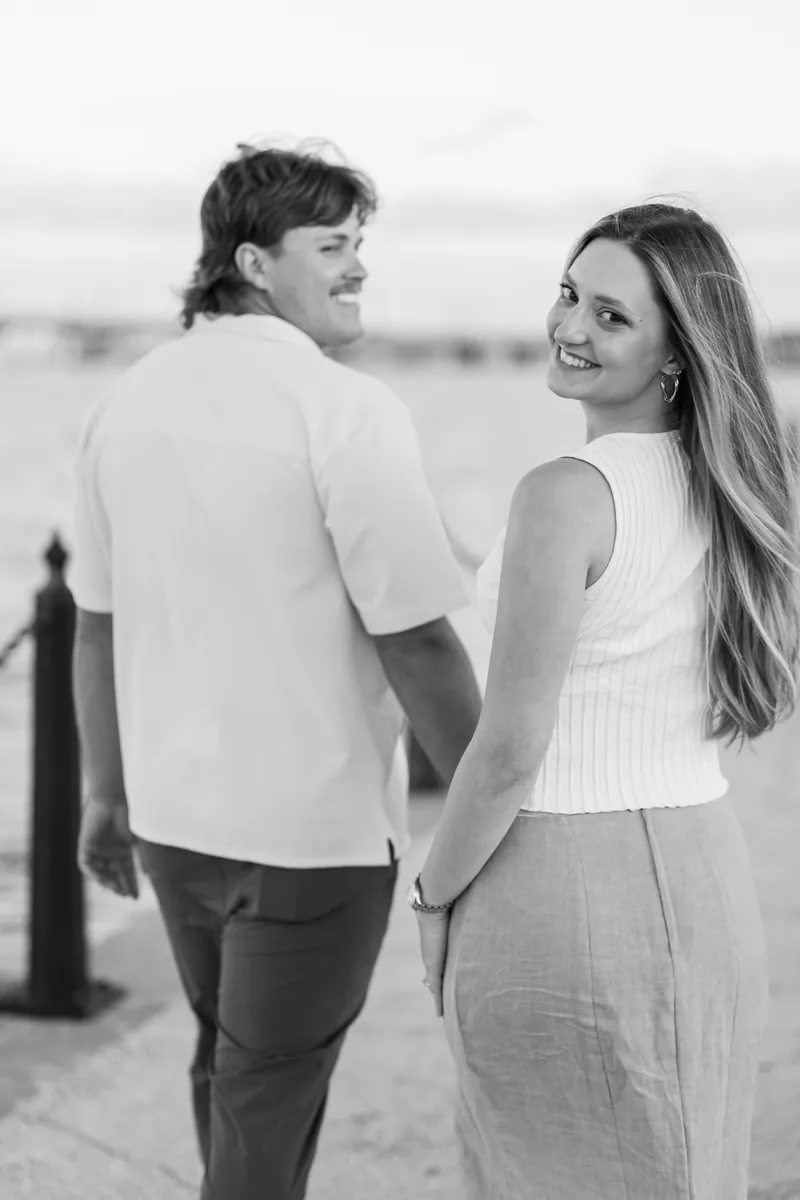 Michael and Katie walk hand-in-hand along a beach in black and white, with wooden posts visible along the shoreline.