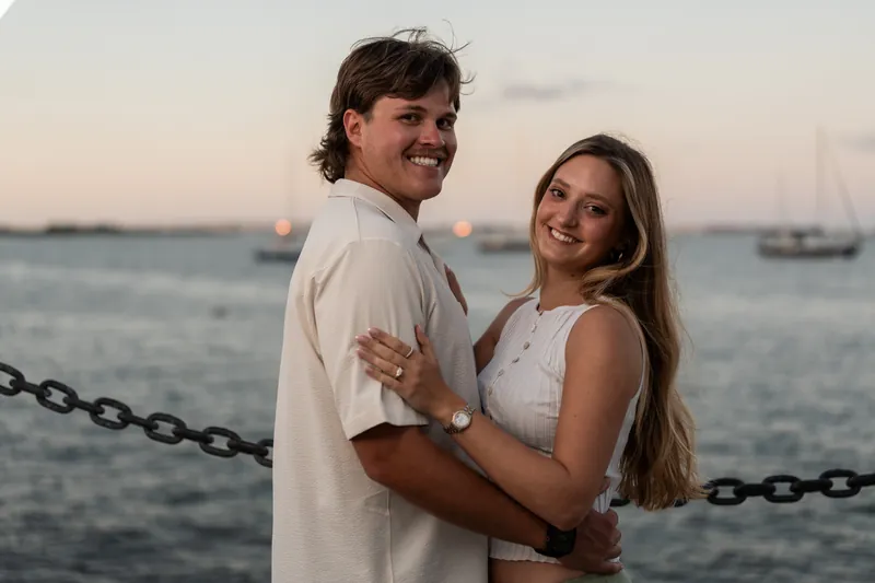 Michael and Katie embrace by a chain railing with boats and calm ocean water behind them at dusk.