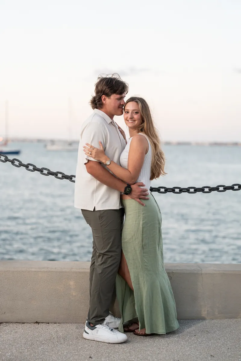 Michael and Katie embrace by a chain railing with the ocean and sunset sky behind them.