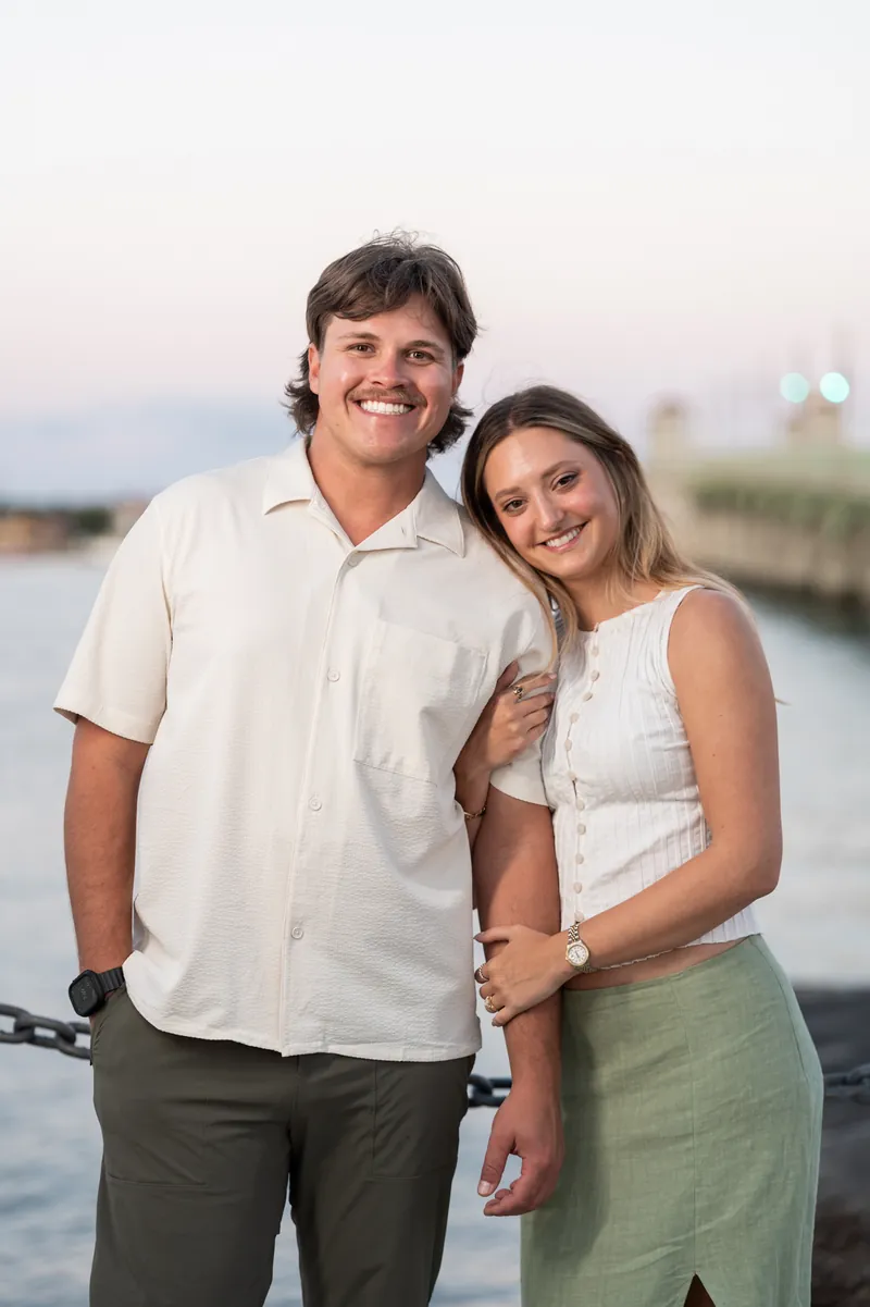 Michael smiles at the camera while Katie leans against his chest on a waterfront pier at dusk.