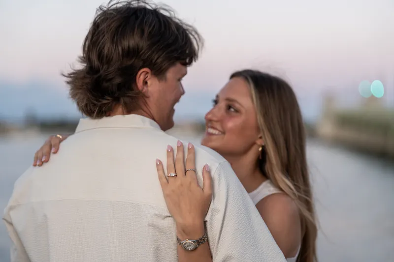 Katie shows her engagement ring to Michael's back as they face the water during golden hour.