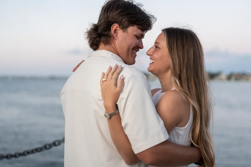 Michael and Katie embrace and laugh together by the ocean with a chain railing behind them.
