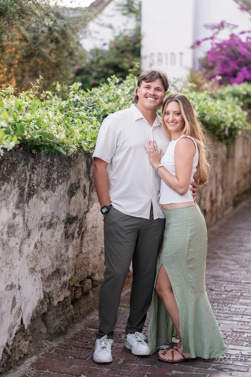 Michael and Katie stand on a garden path lined with flowering bougainvillea, displaying her ring.
