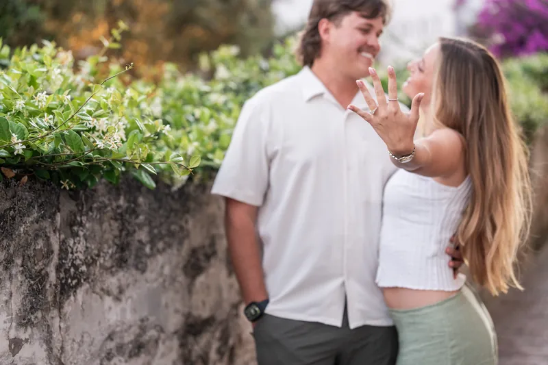 Katie reaches toward Michael's face while standing together in a garden courtyard with blooming flowers.