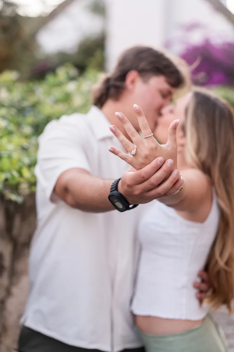 Michael and Katie display their engagement ring while kissing in a garden setting, both wearing white tops.