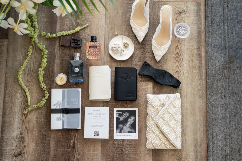 Wedding flat lay with invitation suite, perfume, shoes, vow books, and pearl clutch