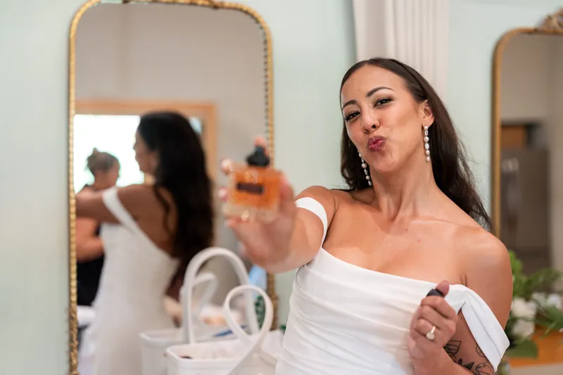 Bride playfully holding perfume bottle toward camera with mirror reflection behind her