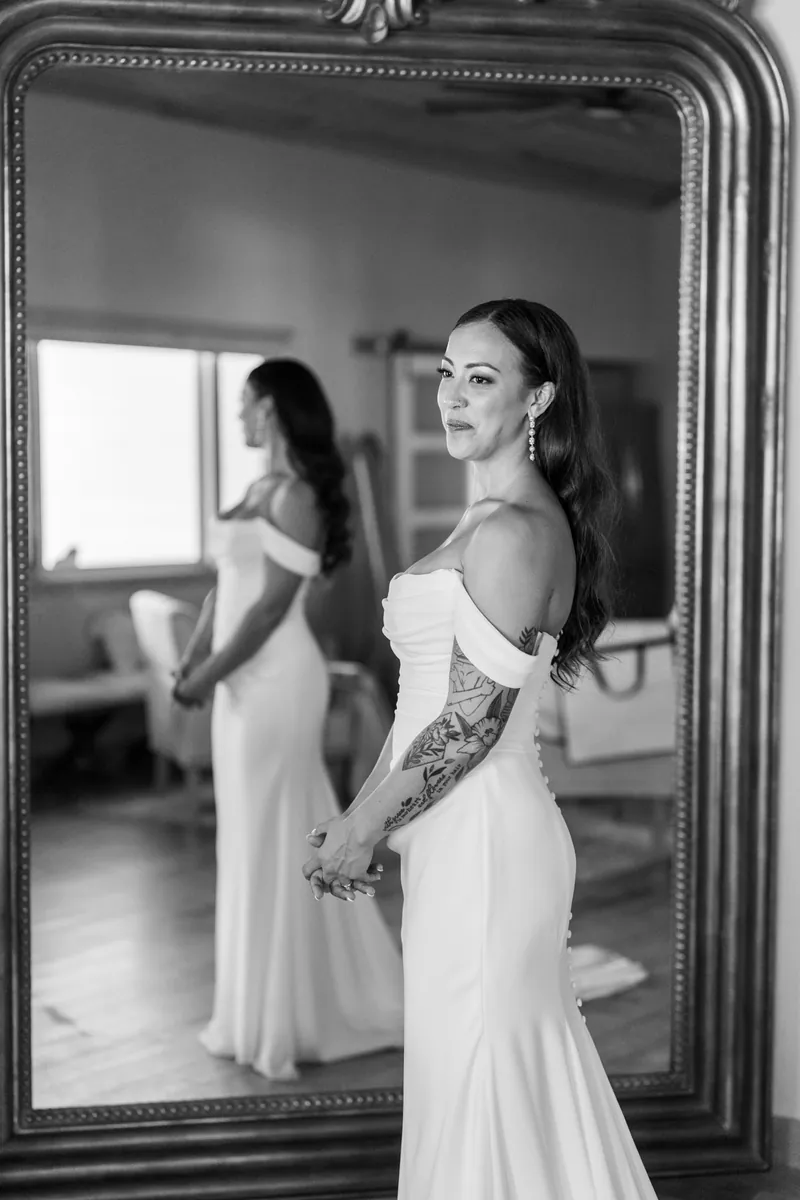 Bride in off-shoulder gown with button-back train standing in front of full length mirror
