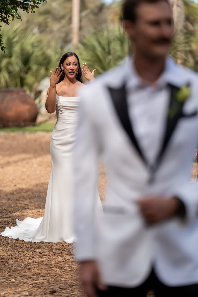 Bride approaching groom from behind during first look among tropical plants and coquina walls