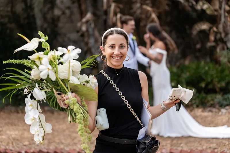 Wedding coordinator carrying the bridal bouquet while couple shares a moment in background