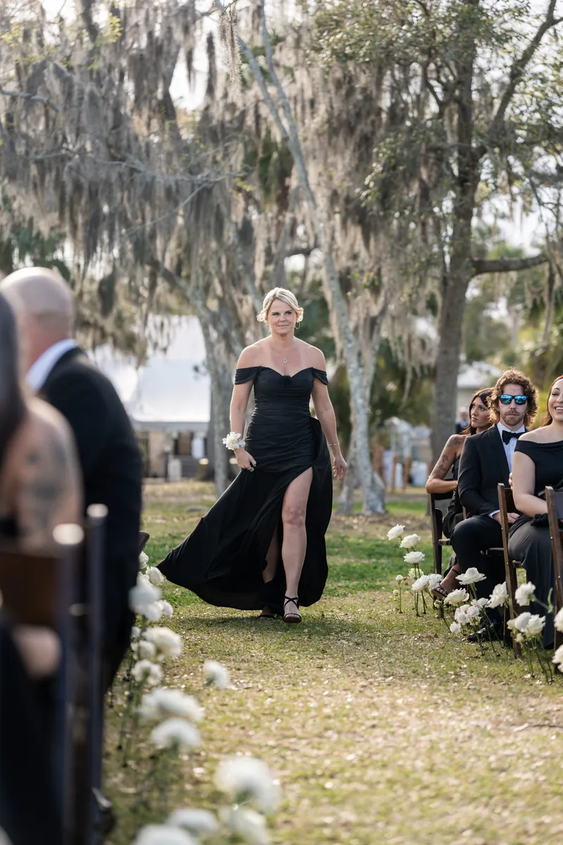 Mother of the bride walking down the aisle in blue floral dress carrying a calla lily