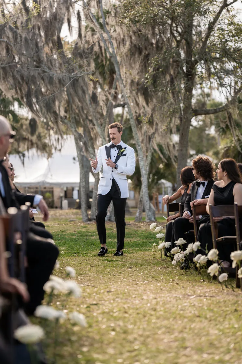 Groom walking down the aisle pointing to guests in his white tuxedo