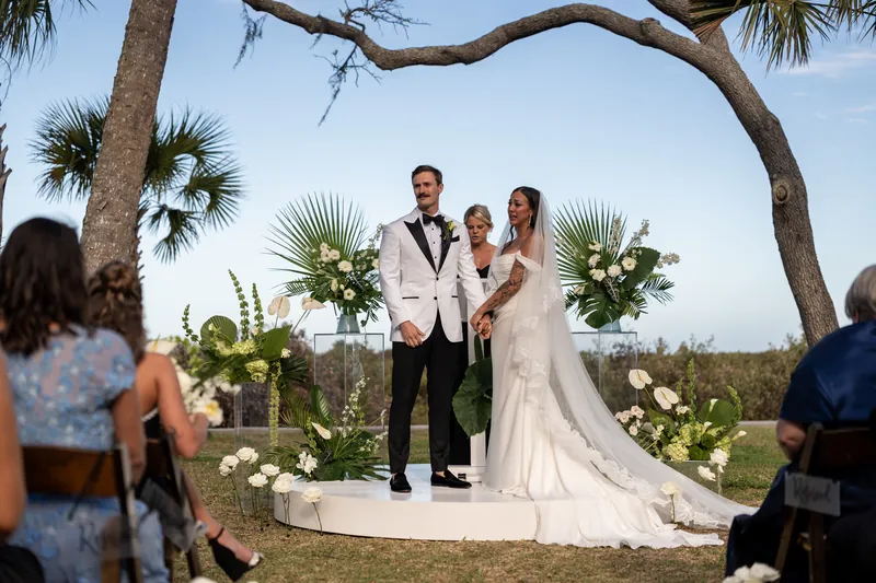 Wide ceremony view with couple on white round platform under palm trees as guests watch