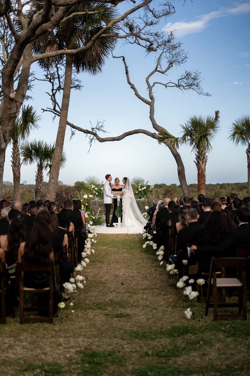 Full ceremony wide shot down the aisle framed by white roses with couple at altar under oaks