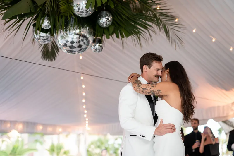 Couple slow dancing under disco balls and palm frond installation at reception