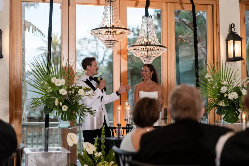 Bryant speaking at microphone while Michelle laughs during reception speech under chandeliers