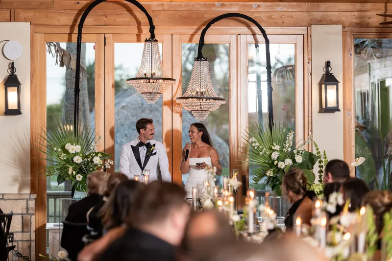 Michelle speaking to guests at reception with Bryant beside her under chandeliers