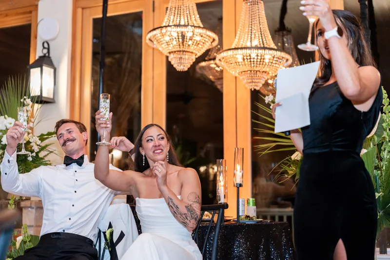 Couple raising champagne glasses during toast at reception with chandeliers