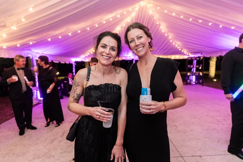 Two guests posing together with drinks under purple lit tent