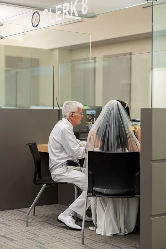 Two people sit at a desk during their marriage ceremony, with one wearing a white dress and veil.
