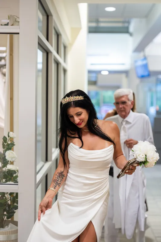 A person in a white ruched dress and tiara walks down a hallway beside their partner holding flowers.