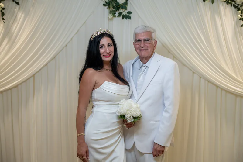 Isury and Mike stand shoulder-to-shoulder in white formal wear, both smiling at the camera with Isury's bouquet visible.