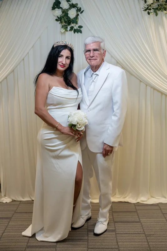 Isury and Mike pose together in their white elopement outfits, with Isury holding a white bouquet and Mike's arm around them.