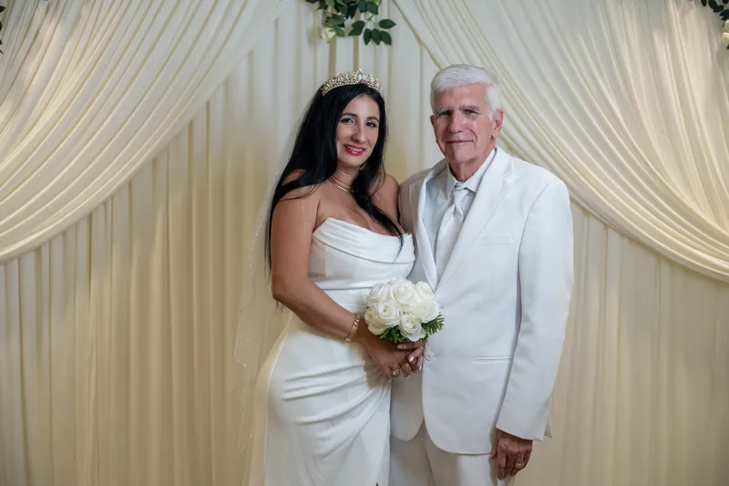 Isury and Mike stand together in white attire holding a white bouquet beneath cream draping at the courthouse.