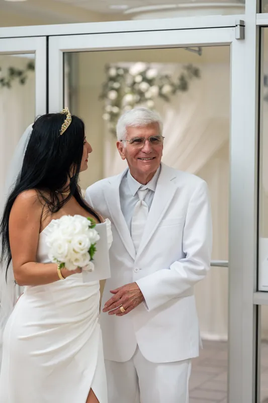 Mike gazes at Isury as they stand in a doorway, Isury holding her white bouquet in her strapless dress.