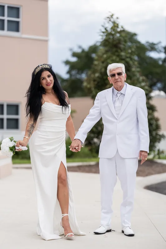 Isury and Mike hold hands while walking across the courthouse plaza, both in coordinating white attire.