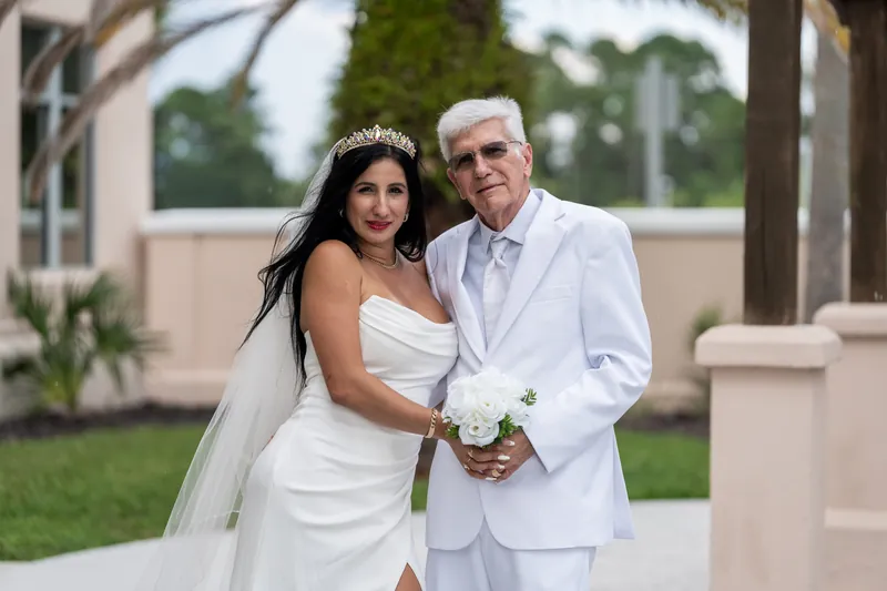 Mike and Isury in white outfits hold a white bouquet outside the St. Johns County Courthouse in St. Augustine.