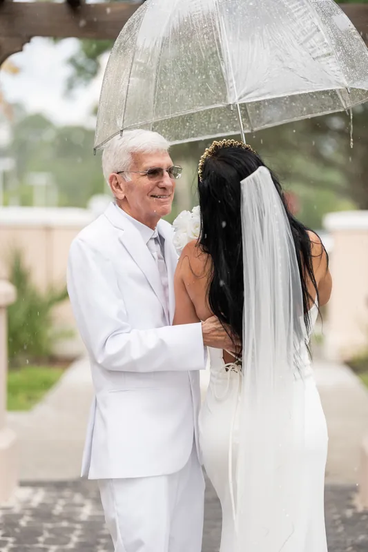 Mike shelters Isury under an umbrella while they stand close together, her veil flowing over his shoulder at the courthouse.